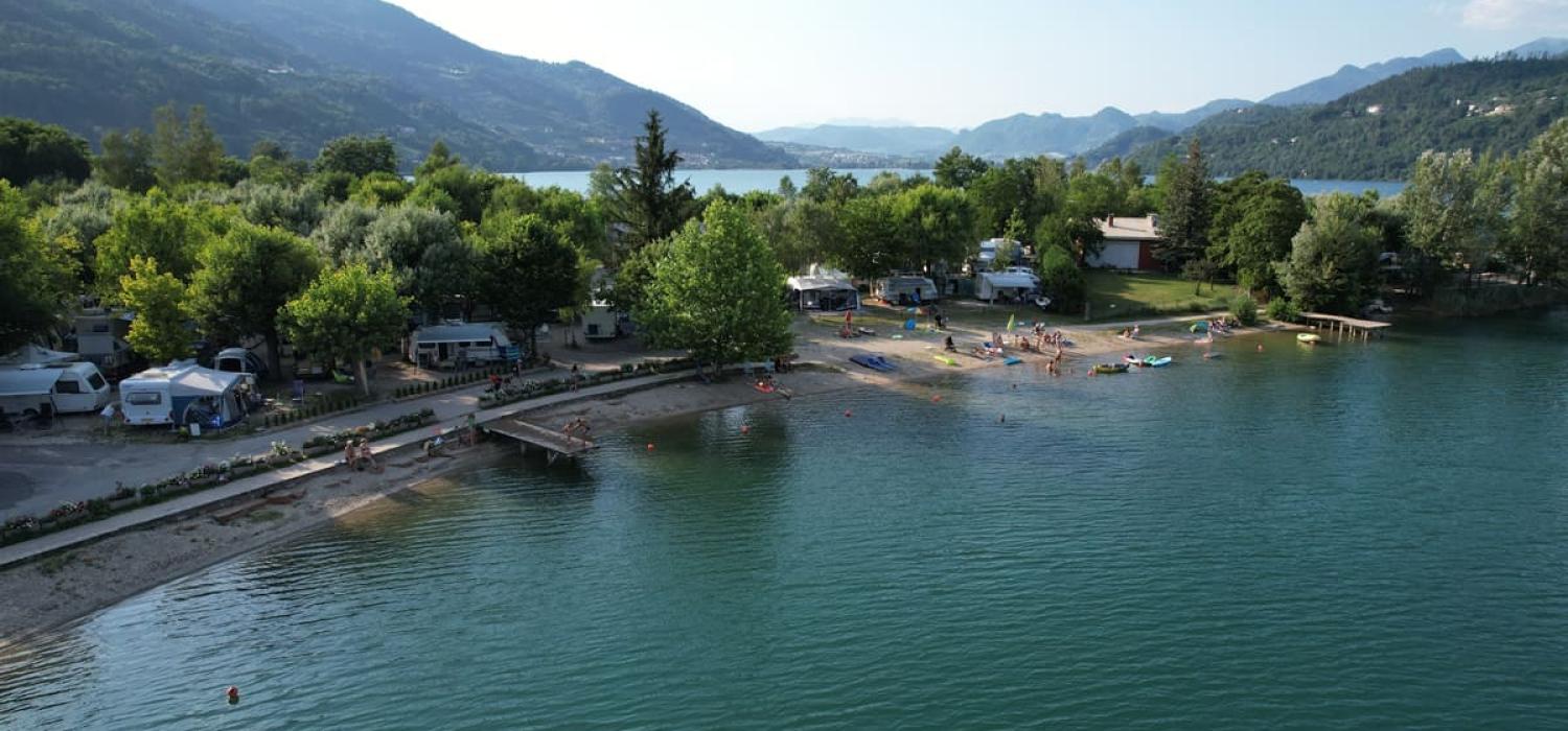 Campeggio sul lago con spiaggia, alberi e montagne sullo sfondo.