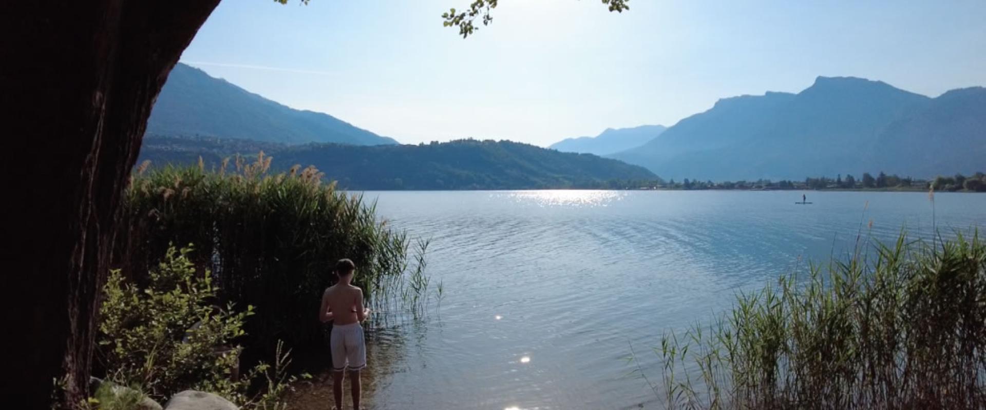 Persona in piedi sul lago, circondata da montagne e vegetazione, giornata serena.