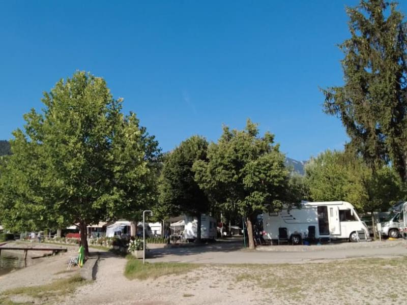 Campeggio sul lago con camper, alberi e una persona che cammina.