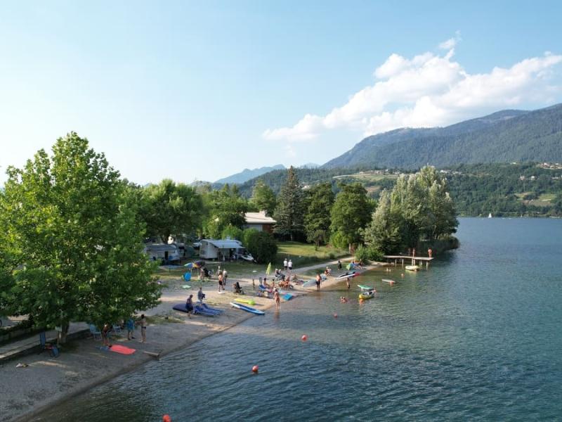 Spiaggia affollata sul lago, circondata da alberi e montagne, giornata estiva serena.