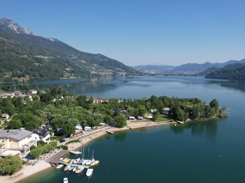 Lago alpino con spiaggia, barche e colline verdi circostanti, sotto un cielo sereno.