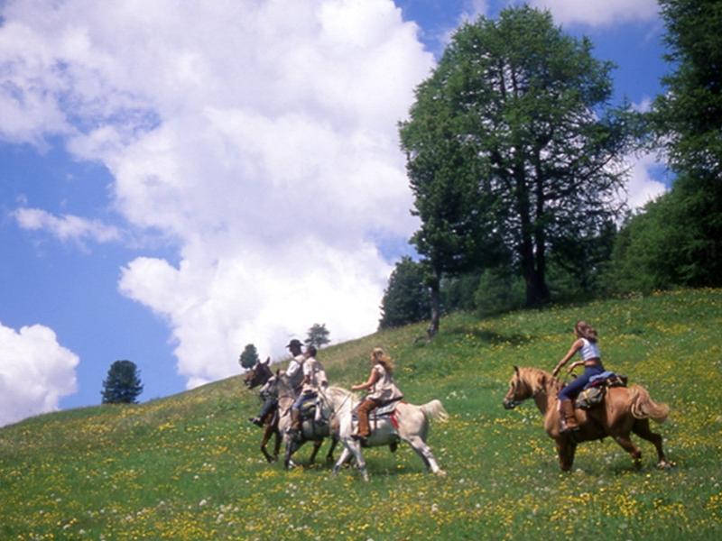 Persone a cavallo attraversano un prato verde sotto un cielo nuvoloso.