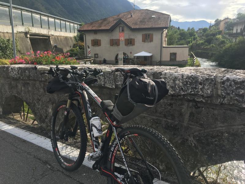 Bicicletta su un ponte di pietra, casa e fiume sullo sfondo montano.