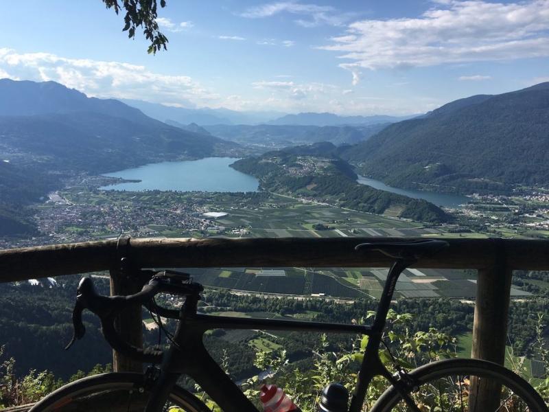 Vista panoramica di un lago con bicicletta in primo piano e montagne sullo sfondo.