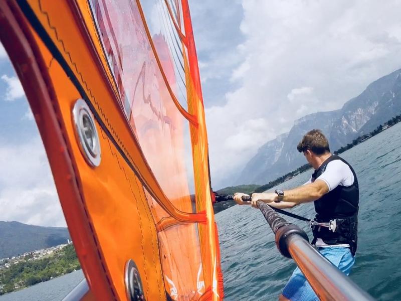 Persona pratica windsurf su lago con montagne sullo sfondo, vela arancione in evidenza.